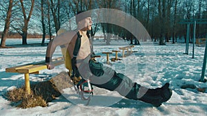 Muscular man with a naked torso and with a drager on his back does reverse push-ups from a bench in a winter park