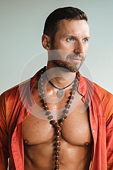A muscular man with a naked torso in orange clothes stands in a room on a light background