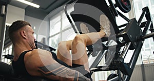 A muscular man performs exercises on a sports training apparatus for leg muscles in a gym, lifting weights