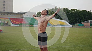 A muscular man stands under drops of water from a sprinkler on a football field with his arms outstretched. Slow motion