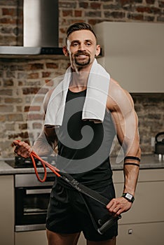 A muscular man with a towel on the shoulders is posing with pull elastic rope