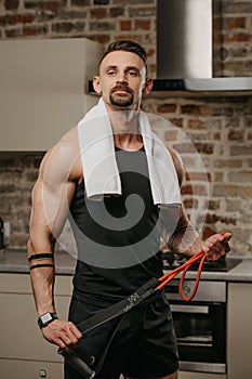 A muscular man with a towel on the shoulders is posing with pull elastic rope