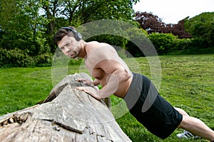 Muscular man, wearing headphones training with pressups against a fallen tree in park