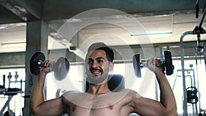 Muscular powerful young man doing shoulders overhead press lifting with dumbbells in fitness gym