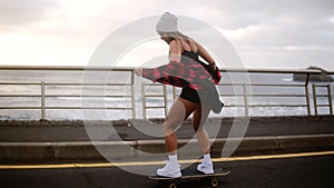 Muscular, pretty hipster woman in plaid coat having fun riding skateboard longboard downhill on beautiful road in slow