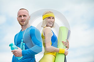 Muscular sport trainer with cheerful smiling young woman holding yoga mat at cloudy sky background. Couple doing sport