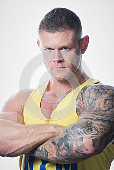 Muscular strong man with blue eyes and tattoo in the yellow tank top on the white background in a gym