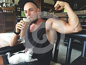 Muscular Strong Vegan Man Showing Biceps and Drinking Iced Coffee Protein Drink. Cute Small Kitten Sitting on Sportsman