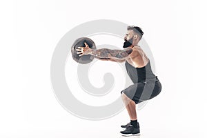 Muscular tattooed bearded male exercising fitness weights Medicine Ball in studio isolated on white background