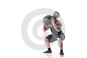 Muscular tattooed bearded male exercising fitness weights Medicine Ball in studio isolated on white background