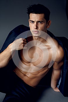 Muscular, young brunette man sitting and posing in fashionable suit on a naked torso,  on grey background.