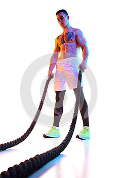 Muscular young man with fit, strong shirtless body standing with battle rope against white studio background in neon