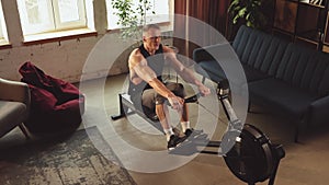 Muscular young man training shirtless with stationary rowing machine, doing exercises at home on daytime. Top view