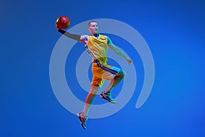 Muscular young man in yellow uniform, basketball player during game, jumping with ball against blue studio background in