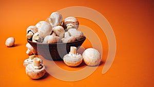 Mushrooms in a wooden bowl on an orange background. The small white champignon in a plate and scattered near it