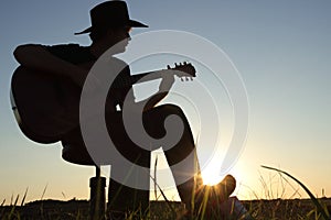 Music concept of black silhouette of man with western hat cowboy and instrument at sunset or sunrise with skyline and grass.