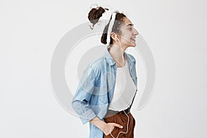 Music, happiness and technology. Lovely stylish girl with hairbun, holds hand in pocket of brown trousers, listens to