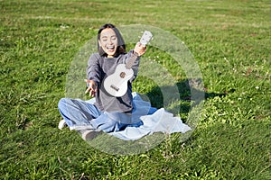 Music and instruments. Portrait of cute asian girl shows her white ukulele, plays in park while sitting relaxed on