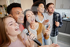 Music for life. Happy multicultured friends singing with microphone while playing karaoke at home, standing in the