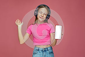 Music mobile app advertisement. Happy little girl child in headphones hold phone with white empty screen mock up