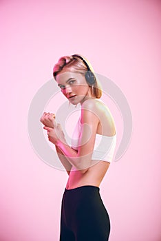 Music is my motivation. Side view of sporty slim blonde woman in headphones listening music while standing against pink