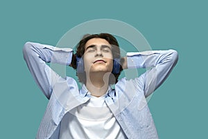 Music relaxation. Portrait of a young beautiful man wearing white t-shirt and blue shirt in headphones threw hands behind his head