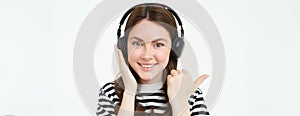 Music store concept. Smiling young woman listening song in headphones, pointing left at copy space, shows advertisement