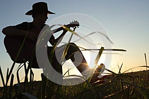 Musical theme scene of silhouette of man with hat and guitar at sunset or sunrise. Skyline of musician with sun and blue sky.