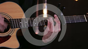 Musican man playing chords on acoustic guitar with a capo on black background.