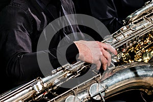 A musician in black clothes sits and holds a baritone saxophone with his right hand. For information on the musical theme.