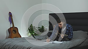 Musician creating new song on electric guitar. Young millennial practicing guitar on bed at modern living room. Man plays lyric mu