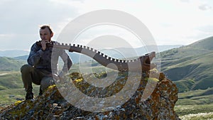 A musician daydreams with an Australian aboriginal musical instrument didgeridoo on a rock ledge on a hillside against