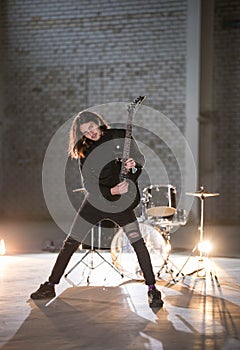 A musician man with long hair playing guitar - looking in the camera - standing on the background of the drum-kit