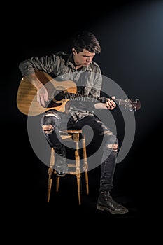 Musician playing acoustic guitar, sitting on high chair, black background with beautiful soft light