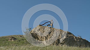 A musician plays the Australian Aboriginal musical instrument Didgeridoo on a rock ledge on a hillside against a blue