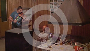 Musician plays violin in wooden room. Woman sitting on chair makes video of show.