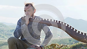The musician pondered with the Australian Aboriginal musical instrument Didgeridoo on a rock ledge on a hillside against