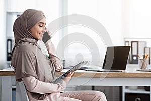 Muslim businesswoman talking on cellphone, holding clipboard, checking documentation in office