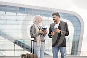 Muslim couple with suitcase, standing outdoors at airport and checking their flight schedule