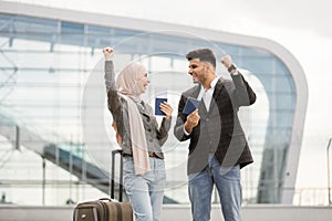 Muslim couple with suitcase, standing outdoors at airport and checking their flight schedule