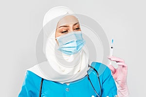 Muslim doctor or nurse in hijab and medical face mask holding a syringe on a gray background. the concept of vaccination of the