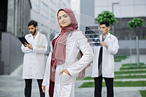 Muslim doctor with stethoscope looks at camera standing outside hospital in front of male colleagues