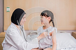 Muslim doctor woman examining heartbeat of girl kid sitting on bed and smile with stethoscope in room at hospital