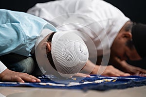 Muslim father and son praying or performing Salah while sitting on Prayer rug and touching head to mihrab or mosque