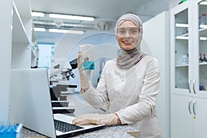 Muslim female chemistry student in hijab sitting at a table in a research laboratory and studying liquid substances