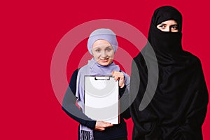 Muslim girls on a red background. A young girl holds a folder, white sheet, smiles and looks at the camera, another