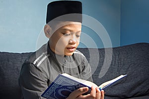 Muslim male student reading the Quran solemnly wearing a skullcap at home, on a blue background
