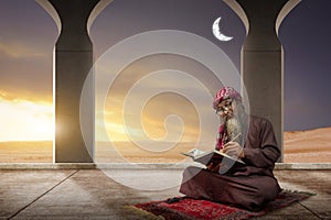 Muslim man with a beard wearing keffiyeh with agal sitting and reading the Quran on the prayer rug