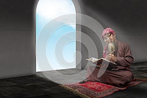 Muslim man with a beard wearing keffiyeh with agal sitting and reading the Quran on the prayer rug