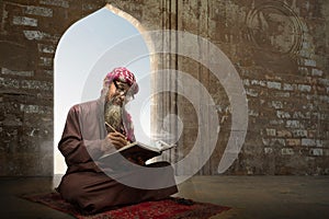 Muslim man with a beard wearing keffiyeh with agal sitting and reading the Quran on the prayer rug